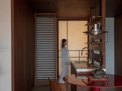 A modern kitchen with a wooden floor, a red dining table, and a variety of furniture and items, lit by natural sunlight