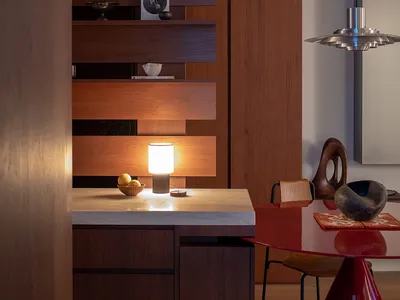 Modern kitchen with wooden cabinets, a red dining table, and dim lighting, featuring a sink and refrigerator, with a warm and inviting atmosphere