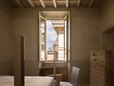 A calm and inviting bedroom with a white color scheme, a bed near the center, a chair by the window, and natural light from the window providing a view of the city