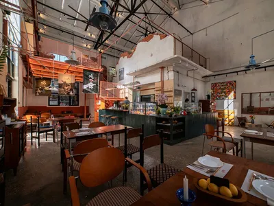 Cozy restaurant interior with tables, chairs, and a bar, featuring potted plants, vases, and a bowl of fruit, with a mix of natural and artificial lighting