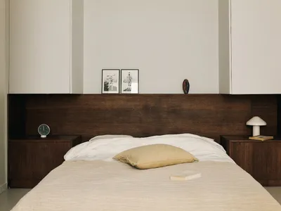 A calm and inviting bedroom with a white comforter, a tan pillow, a nightstand with a lamp, and a dresser in the corner, set against a clean and well-organized white-walled space