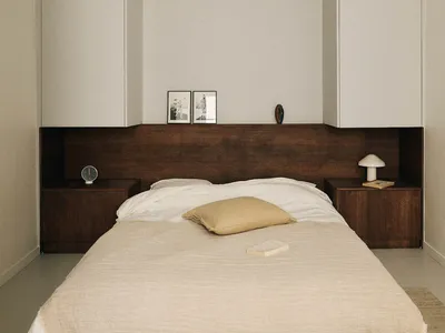 A well-lit bedroom with a bed, nightstand, and dresser, featuring a white comforter, brown pillow, and wooden furniture