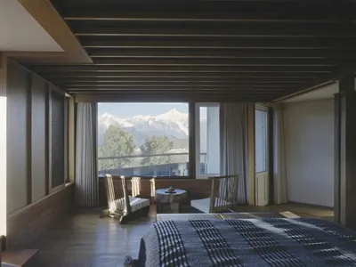 Cozy bedroom with a wooden floor, wooden ceiling, a bed with a plaid blanket, a chair, and a dining table in the corner, with natural light coming in through the window