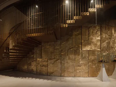 A dimly lit room with a brown stone staircase and wooden railing, set against a large stone wall