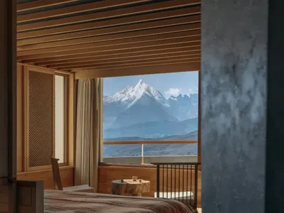 A bedroom with a bed, chair, and window, featuring wood and fabric materials in a cozy and inviting atmosphere with natural light from the window