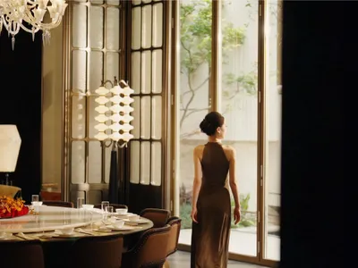 A woman stands in front of a window in a large, elegant dining room with a wooden dining table and chairs, surrounded by wine glasses