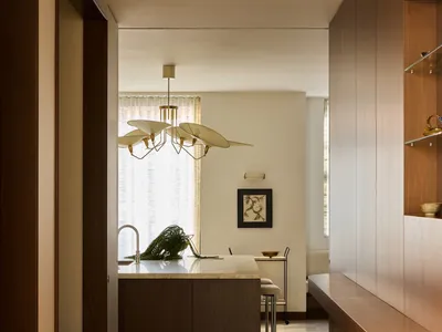 A modern kitchen with wooden floors, cabinets, and a dining table, lit by natural light from the windows, with a potted plant and chairs in the space