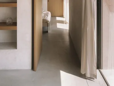 A cozy bedroom with a wooden floor, a white wall, a bed, a bookshelf, and a vase on the bookshelf, with natural light entering through an open door