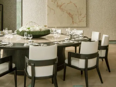 Dining room with a round table set for a meal, surrounded by chairs, a chandelier, and natural light from the windows