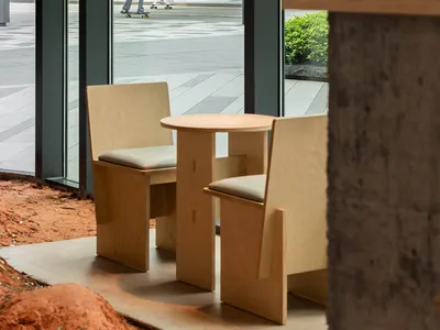 A modern restaurant with a wooden table and glass top, two chairs, and a large window allowing natural light to enter, creating an inviting and comfortable atmosphere for guests