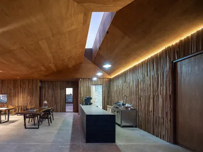 A dimly lit restaurant dining area with wooden walls, a wooden floor, and a dining table surrounded by chairs, a TV on the wall, and a counter with a microwave and sink
