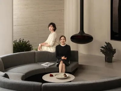 兩位女性坐在沙發上，現代明亮的客廳內有餐桌與盆栽，周圍使用白色素材