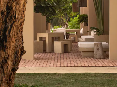 View from a garden into a minimalist outdoor living courtyard featuring geometric concrete tables, stools, and daybeds with white cushions on terracotta brick flooring, surrounded by lush tropical plants and hanging wooden lanterns.