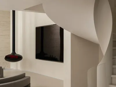 Modern minimalist interior featuring a sculptural curved white staircase, floating black pendant light, built-in TV, and curved grey sofa against textured concrete walls.