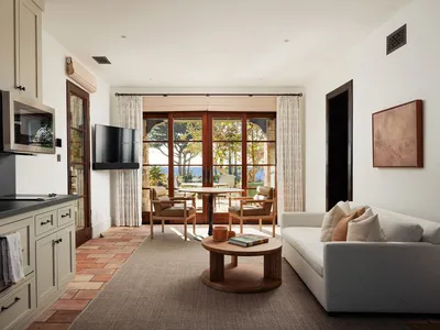 Bright open-plan living room featuring terracotta floor tiles, a light gray sofa, round oak coffee table, two leather armchairs, wall-mounted TV, and large glass doors opening to a coastal terrace with ocean view.