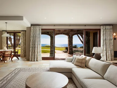 Bright coastal living room featuring a large L-shaped sectional sofa, round wooden coffee table, patterned area rug, and expansive ocean view through three brick arches leading to a patio.