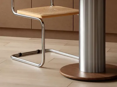 A light wood cane chair with chrome frame sits beside a round walnut pedestal dining table featuring a stainless steel column base, with a Basquiat book and walnuts on the tabletop.