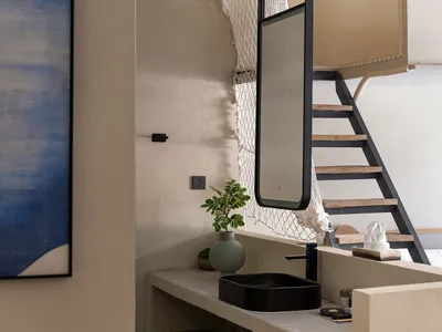 Modern minimalist bathroom vanity made of concrete with a black rectangular vessel sink, black faucet, round mirror, and potted plant against a textured beige wall with a staircase in the background.