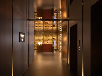 Modern luxury hotel corridor with dark paneling and vertical light strips leading to a brightly lit multi-level wine cellar with a person walking in the background.