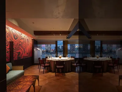 Luxurious private dining room featuring a large round table, red marble accent walls, walnut herringbone flooring, and floor-to-ceiling windows overlooking a harbor city skyline at dusk.
