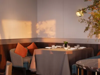 Modern restaurant dining area featuring a round table with grey tablecloth, built-in banquette with orange cushions, walnut wood ceiling beams, and warm ambient lighting casting soft shadows on the wall.
