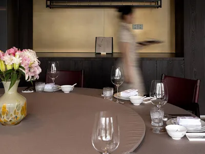 Modern dining room with a large curved table set with glassware and flowers, featuring a brass wall with black floating shelves holding wine glasses and liquor bottles, dark wood paneling, and burgundy leather chairs.