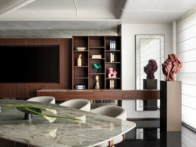 Modern luxury dining room featuring a large green marble dining table, light beige upholstered chairs, a walnut media wall with built-in shelving, and dramatic red wood sculptures on pedestals.