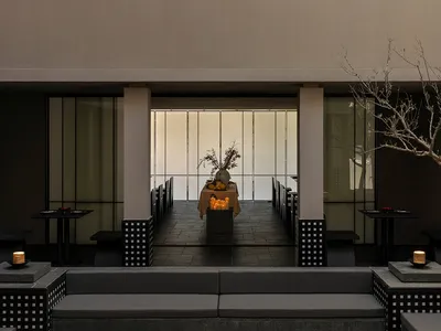 Modern minimalist outdoor dining courtyard at dusk featuring concrete architecture, dark furniture, candlelit tables, and a central dining setup with large windows in the background.