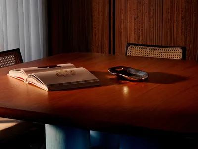 Close-up of a modern wooden dining table with a thick, rich brown tabletop and distinctive bright blue cylindrical and spherical legs, an open book and metallic bowl resting on the surface, with cane-back chairs visible in the background.