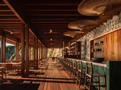 Long open-air wooden veranda with teak flooring and ceiling, featuring a stone wall, green tiled bar counter, woven rattan pendant lamps, and rows of wooden tables and chairs overlooking a dramatic coastal cliff.