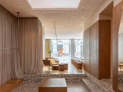 Modern minimalist boutique interior featuring irregular terrazzo flooring and steps, warm oak wood paneling, neutral linen curtains, and contemporary seating areas with wooden tables and leather chairs.