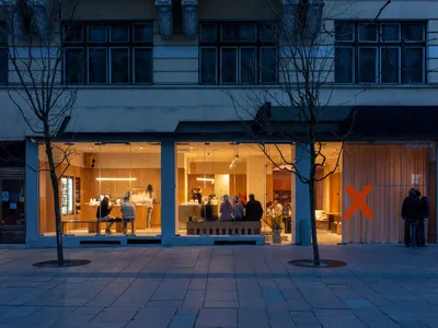Night view of a modern minimalist cafe named Ukyo X with floor-to-ceiling glass windows revealing warm wooden interiors and patrons inside, located on a paved European street with bare trees.