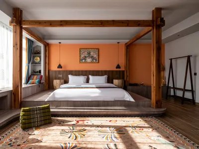Bright bohemian bedroom featuring a large wooden four-poster bed, burnt orange accent wall, white linens, and colorful patterned rug with sunlight casting strong shadows.