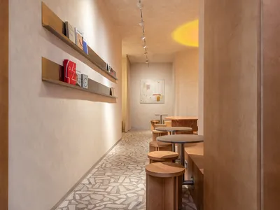 Narrow minimalist cafe interior with terrazzo tile floor, wooden bar stools, brass floating shelves displaying books, and warm ambient lighting.