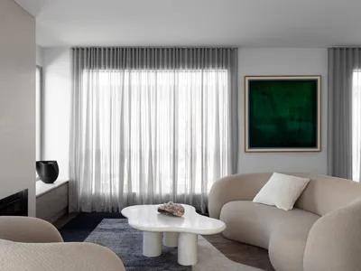 Contemporary living room with beige boucle curved sofa, white irregular marble coffee table, dark blue rug, sheer curtains, and large green abstract painting.