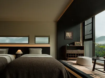 Modern bedroom with two beds covered in brown textured throws, dark wood headboard with backlighting, wooden console with speaker, and large windows revealing a foggy mountain landscape.
