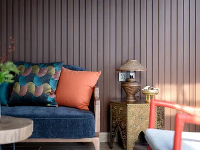 Blue velvet sofa with colorful patterned and solid orange cushions, placed next to an ornate brass side table with a woven lamp against a dark wood slatted wall.