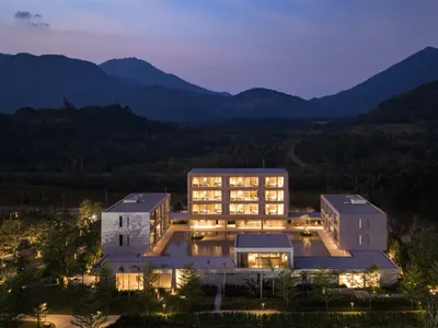 鳥瞰現代多棟度假酒店建築群，中央設有倒影池，在暮光中全亮照明，周圍環繞森林山脈。