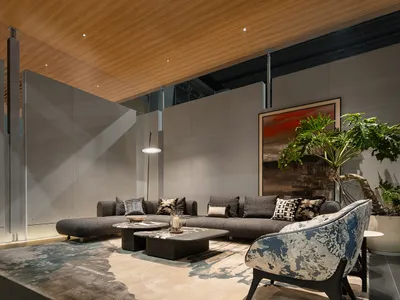 Modern living room with grey L-shaped sectional sofa, blue and white patterned accent chair, concrete walls, wooden ceiling, and large potted plant.
