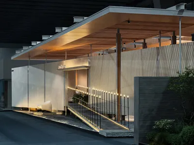 現代極簡商業建築入口夜景，包含混凝土牆面、垂直木板、懸挑溫暖木質雨棚、發光坡道欄杆，以及白色雕塑感座椅。