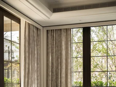 A contemporary living room corner featuring a blue and white checkered upholstered armchair, a textured gold cylindrical side table, a large marble coffee table with a floral arrangement in a brass vase, and floor-to-ceiling windows with neutral curtains.