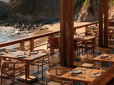 戶外用餐區，柚木餐桌搭配藤編座椅，位於木棧板露臺上，俯瞰沙灘、岩石峭壁與大海，晴空萬里。