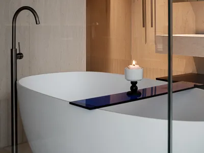 Modern minimalist bathroom featuring a white freestanding oval bathtub, tall black floor-mounted faucet, travertine stone walls, and a lit candle on the tub edge.