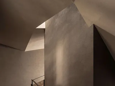Modern concrete staircase with dark wood handrail ascending under angular geometric ceiling planes that create strong directional shadows.