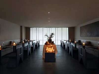 Interior view of a minimalist fine dining restaurant featuring dark stone floors and walls, built-in banquette seating, angular dark wood tables and chairs, and numerous lit candles creating warm ambient lighting, with large floor-to-ceiling windows at the far end.