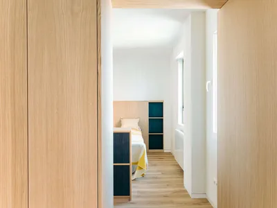 View from a doorway into a narrow modern bedroom with light oak wood paneling, built-in teal storage units, and a bed with yellow and white bedding.