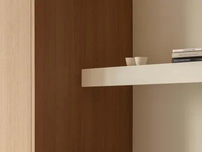 Close-up of a modern dining area featuring warm walnut lower cabinets with round silver knobs, a stainless steel countertop, light oak upper cabinetry, and a cane-backed dining chair in the foreground.