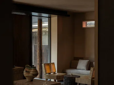 Modern minimalist lounge with raw stone blocks, woven rattan chairs, dark tiled floor, and a glowing square skylight in an earthy-toned interior.