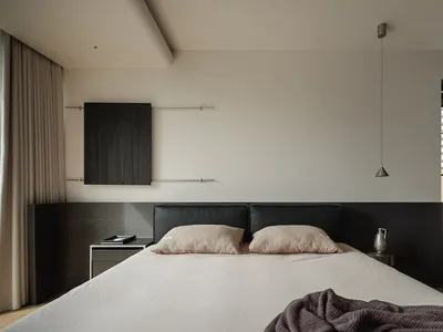 Modern minimalist bedroom with a large bed dressed in white linen and beige textured pillows, set against a dark wood floating headboard panel. A single pendant light hangs above, with a black wood wall cabinet mounted on the left wall.