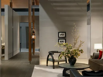 Modern minimalist living room featuring exposed concrete walls, a dark L-shaped sofa, a round glass coffee table, a sculptural black chair, and a large dramatic floral arrangement in a black vase.